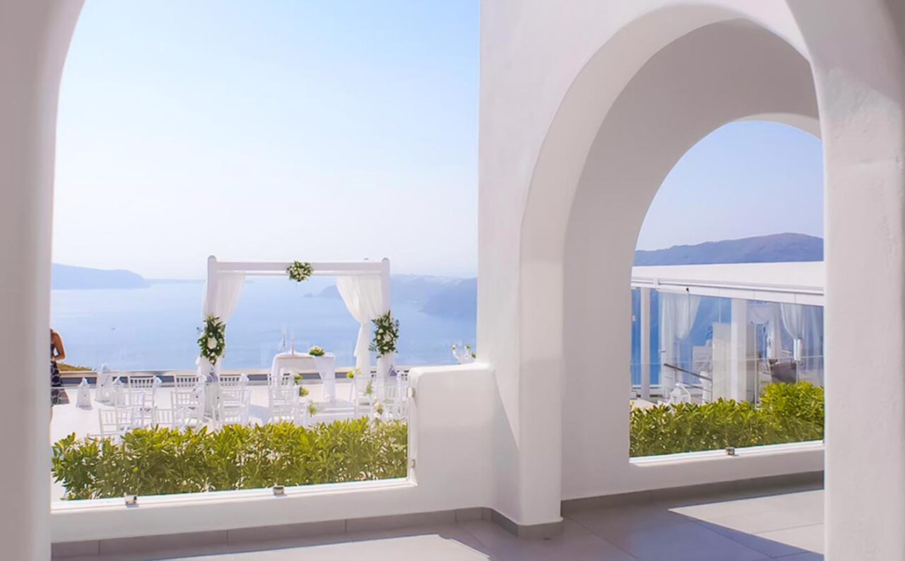 Μίνιμαλ στολισμός γαμήλιας τελετής με θέα την καλντέρα, μέσα από τις χαρακτηριστικές καμάρες ενός χώρου στη Σαντορίνη.