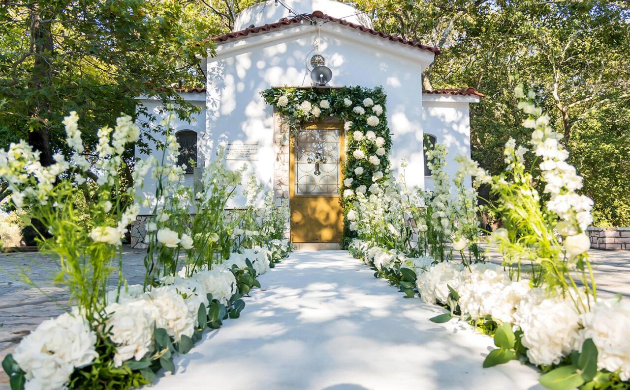 A serene white chapel adorned with lush flowers, designed for an elegant wedding ceremony in Greece.
