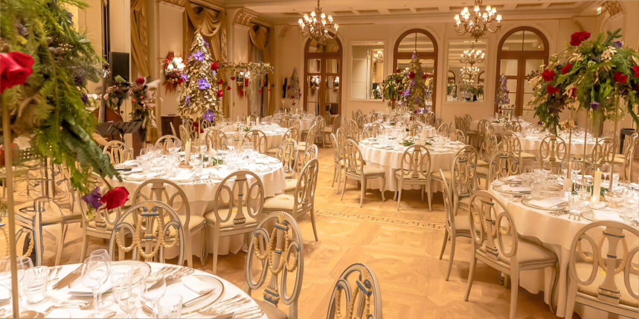 The ballroom glows with luxury as Rogdaki Events decorates round tables with candlelight, gold accents, and grand floral arrangements for the gala dinner and concert