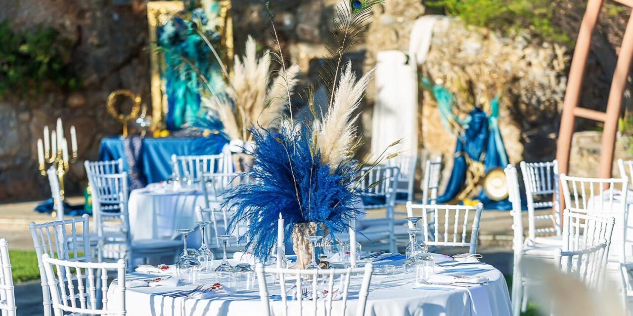 Ancient Greek Decoration in an Amphitheater, by Rogdaki Events — Elegant white tables adorned with blue accents, nestled under mountains and theatrical arches