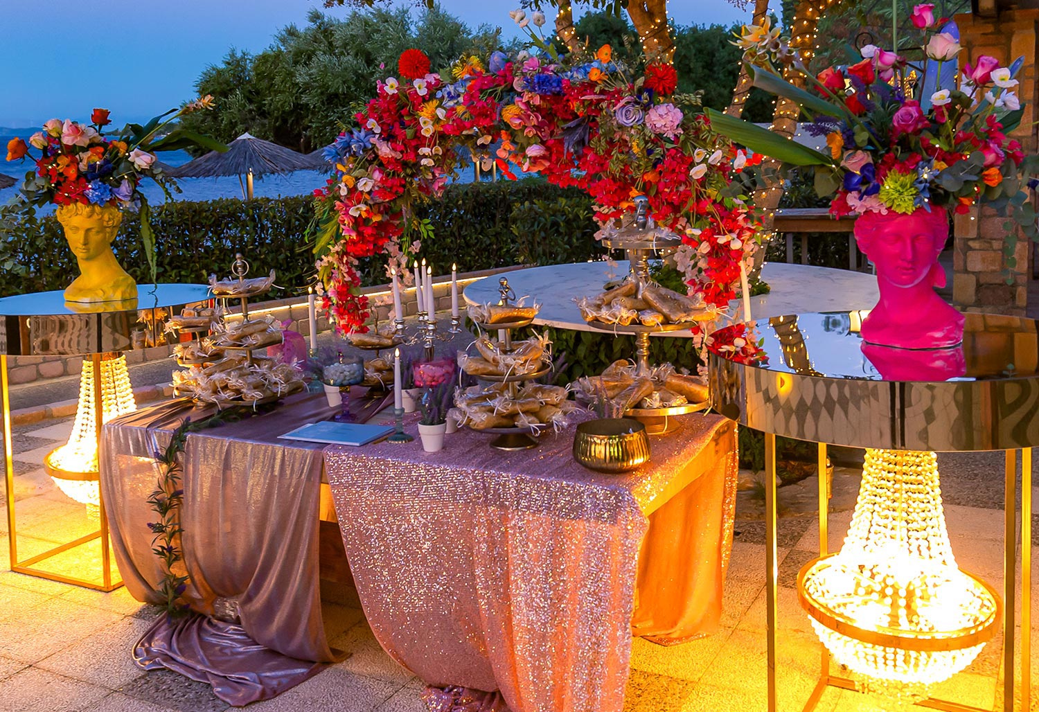 Chic & Colorful Γάμος στο Κτήμα Παναγιωτόπουλου 48 Rogdaki Events, ένας φωτισμένος μπουφές γλυκών με λαμπερούς κρυστάλλινους πολυελαίους και πολύχρωμες ανθοστήλες την ώρα του δειλινού.